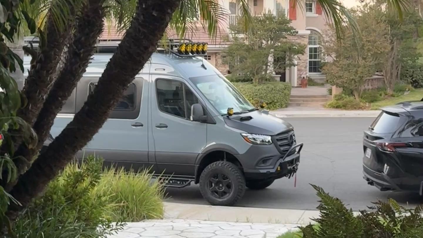Lifted 4x4 Mercedes-Benz Sprinter van with a roof rack and light bar parked on a residential street